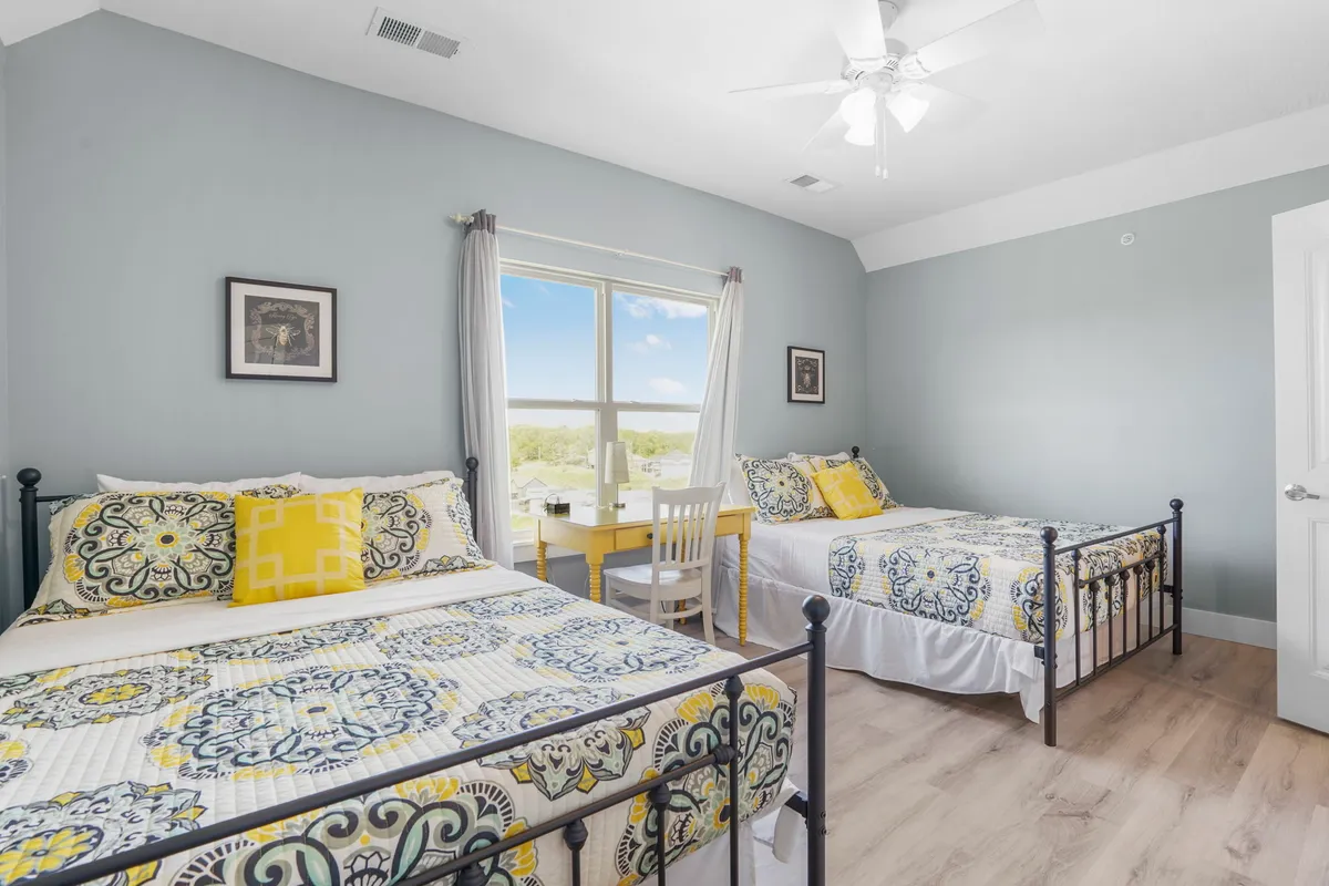 A cheerful bedroom featuring two beds with colorful bedding, a large window offering sunlight and views, and a small table and chairs for reading or quiet activities. The atmosphere is both playful and restful, suitable for families or friends.