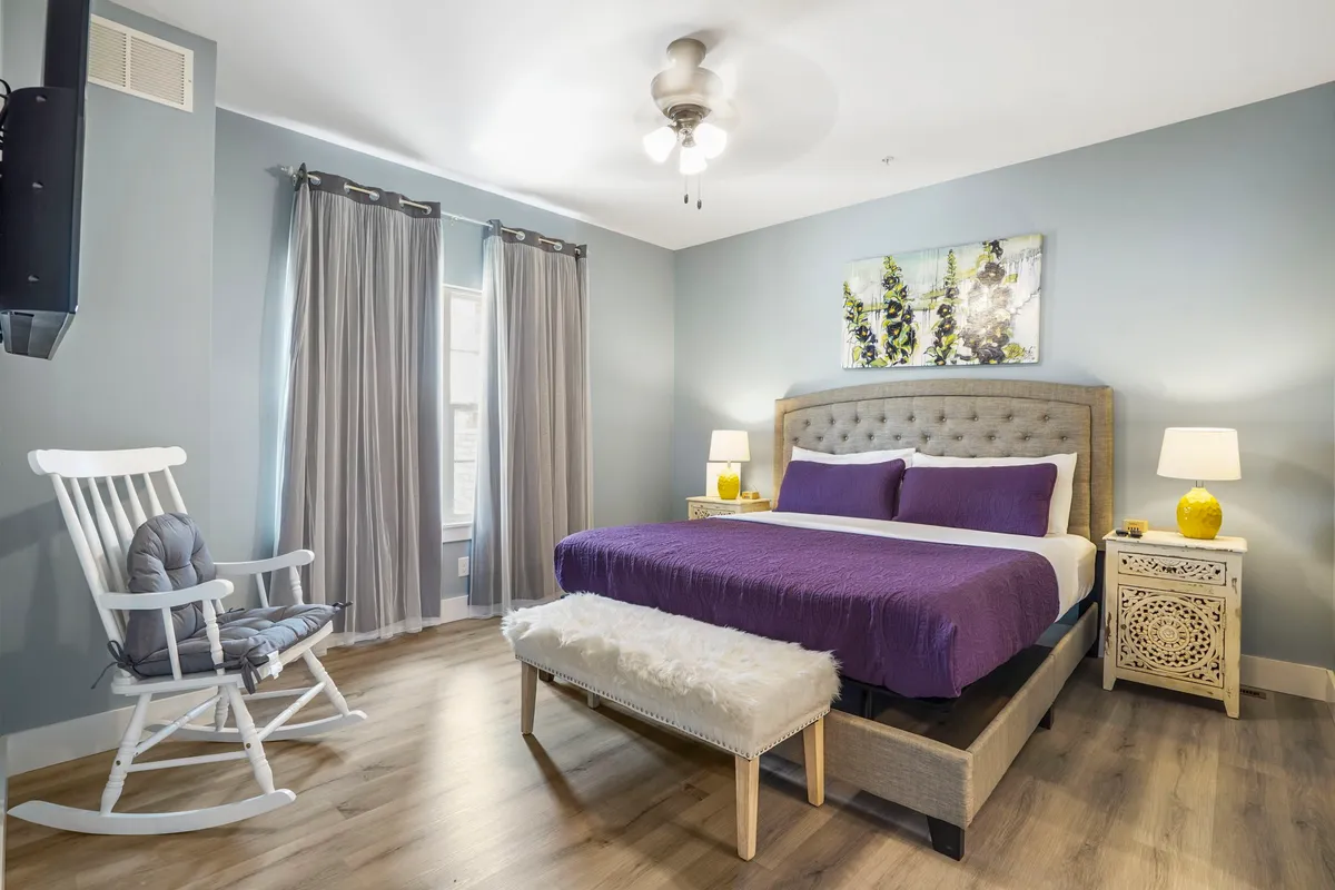 A cozy bedroom with a bed dressed in purple bedding, a comfortable rocking chair, decorative nightstands, and soothing artwork above the bed. Soft natural light fills the space, highlighting its peaceful ambiance and inviting guests to unwind.