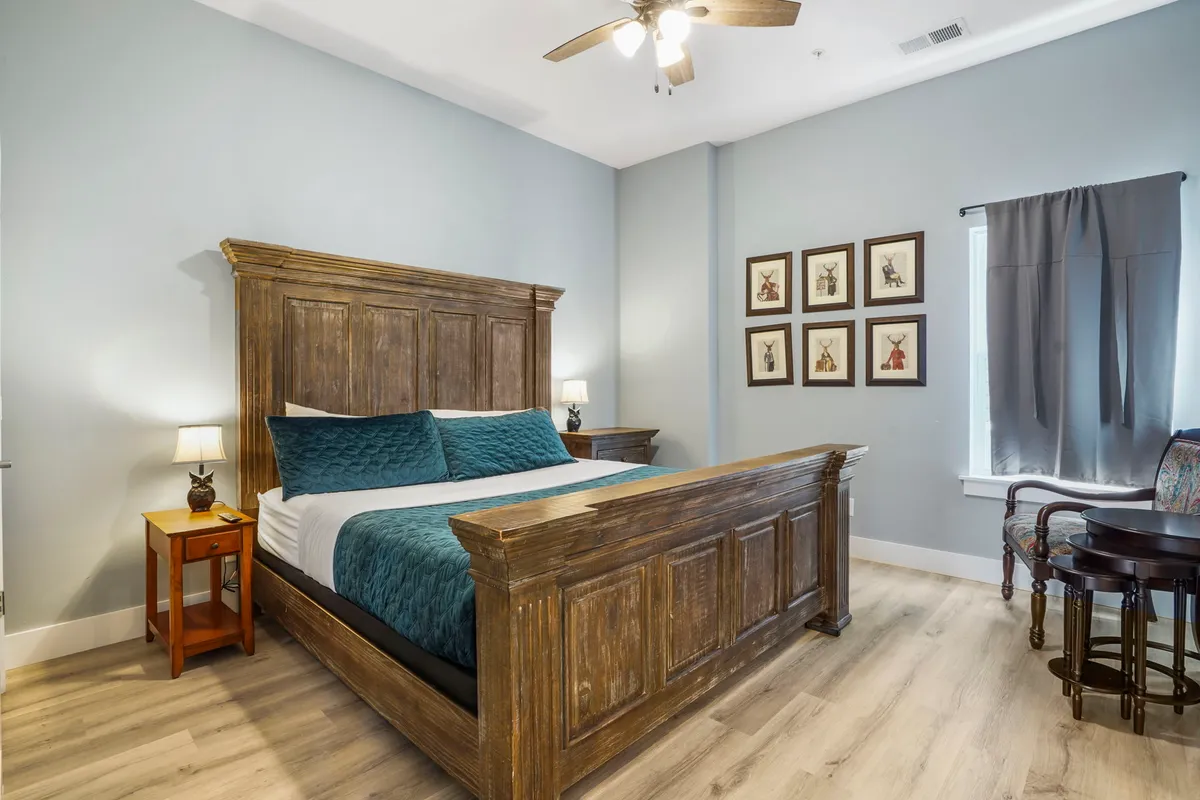 A serene bedroom featuring a wooden bed frame with textured blue pillows, framed art on the wall, and a nightstand with a lamp. The room exudes calm and is bathed in gentle natural light, creating an inviting atmosphere for relaxation or reading.