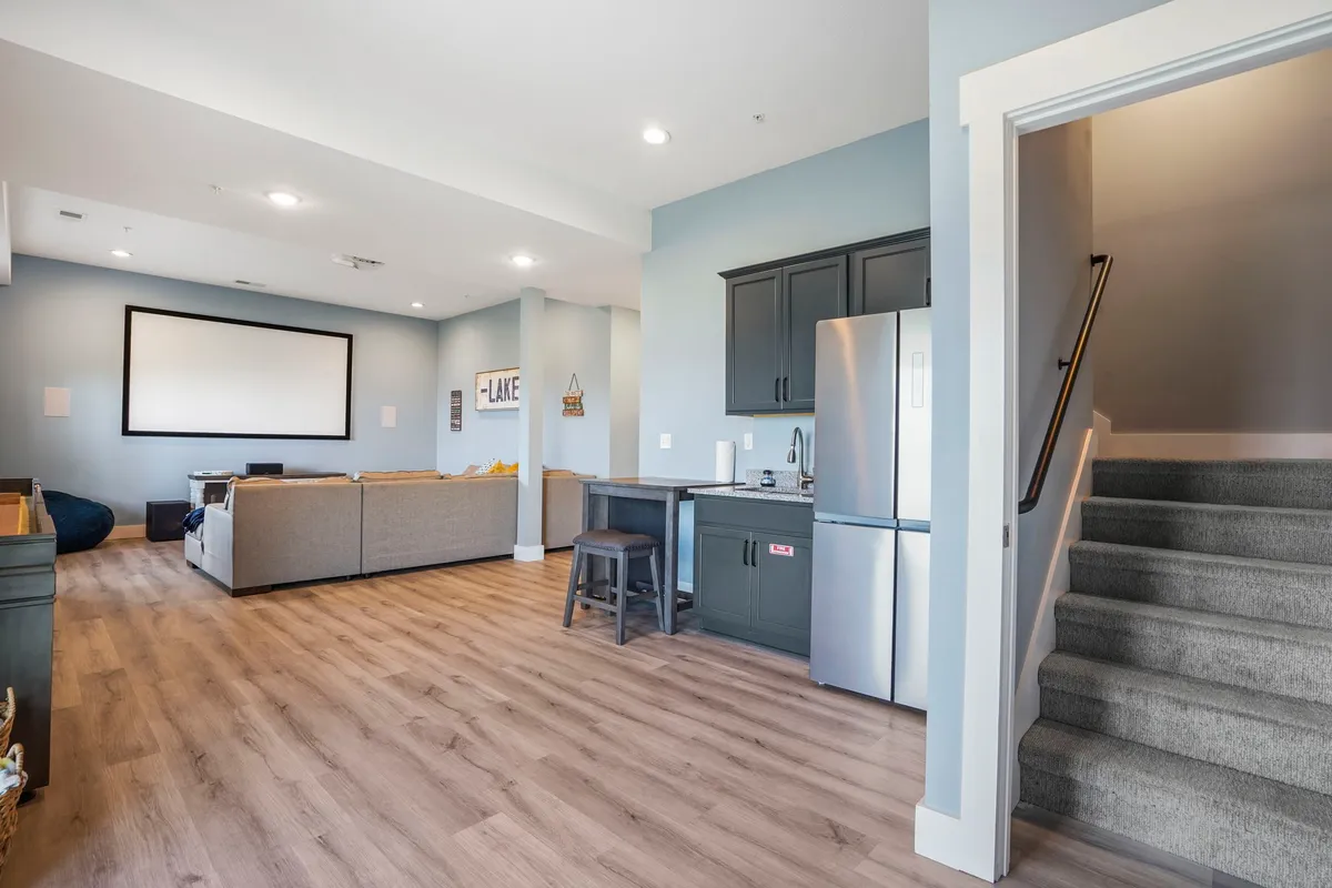 A welcoming entertainment room with a large screen suitable for movies or presentations, a comfortable seating area, and a kitchenette visible in the background—complete with an extra fridge. The relaxed vibe encourages both group fun and laid-back evenings.