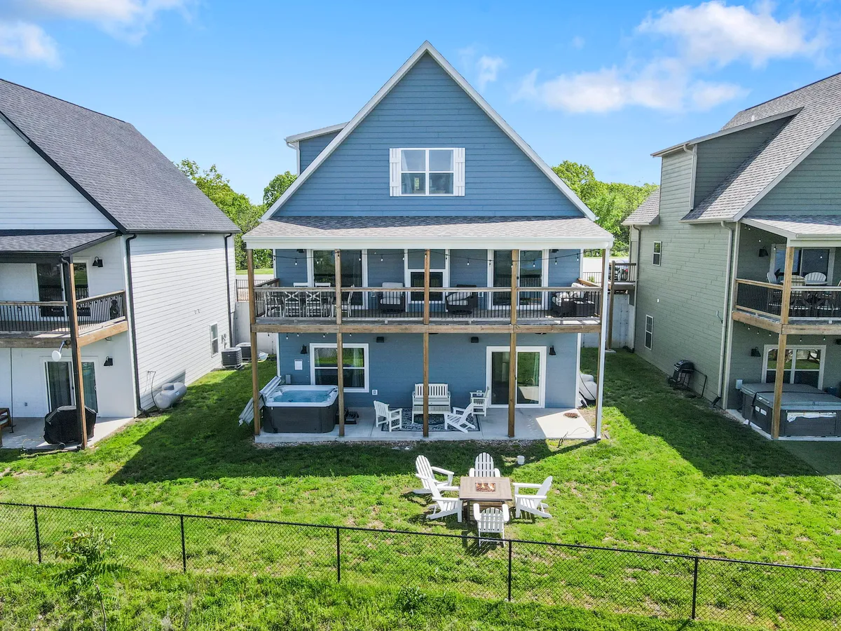 This exterior shot captures a two-story house with blue siding, a spacious deck perfect for morning coffee, a hot tub, and a fire pit area surrounded by chairs. The scene is lively and social, ideal for gatherings and enjoying both sunrise and sunset views.