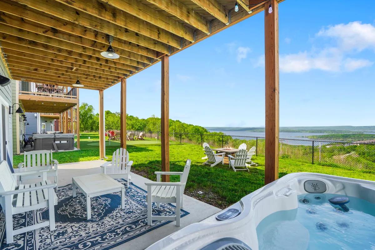 The image shows a spacious patio overlooking a sparkling lake, framed by lush greenery. In the foreground, a steaming private hot tub invites relaxation, while a stylish outdoor seating area encourages gatherings with friends or family. The sun is setting over the water, bathing the scene in a warm, golden glow.