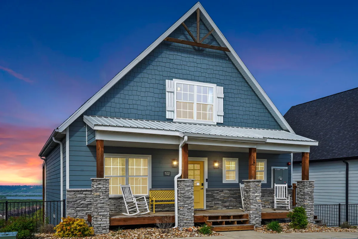 The image showcases the front porch of the cottage, featuring a yellow accent door, rock columns, and shuttered windows. The inviting porch offers a cozy spot for relaxing or gathering with friends and family, with hints of the home’s welcoming atmosphere.