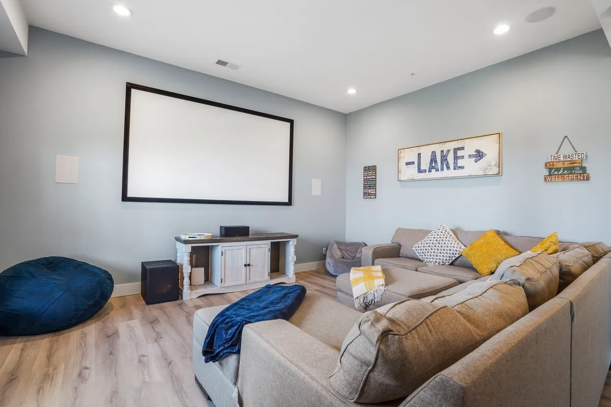 A cozy lower-level room featuring a large empty wall designed for projection, a spacious and comfortable sectional sofa, lake-themed decor, and a bean bag chair. The setup radiates a relaxed, family-friendly atmosphere perfect for watching movies or playing games together.