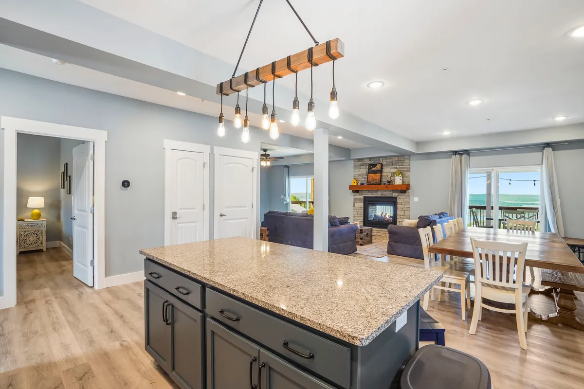 The image highlights a beautifully integrated kitchen, dining, and living area. The kitchen island transitions seamlessly to a dining table, both positioned to take advantage of the expansive lake views through large windows. A fireplace adds to the cozy ambience, making the entire space ideal for entertaining or family meals.