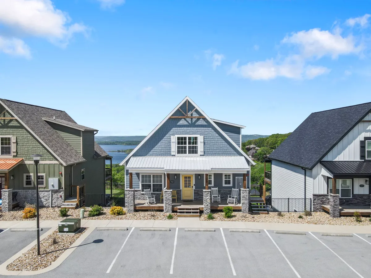 This image features a charming A-frame house with blue siding, a welcoming front porch furnished with rocking chairs, and lush greenery all around. The porch overlooks a serene water view, providing a direct visual connection to the lake and the surrounding outdoors.