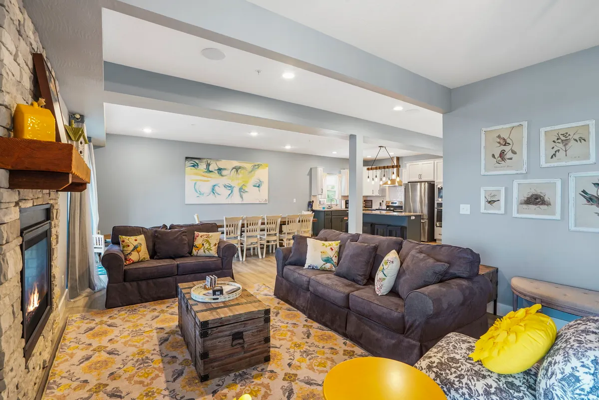 A spacious, sunlit living and dining area features a double-sided fireplace as a central focal point. Plush sofas are arranged to encourage conversation and relaxation, while a large dining table sits nearby, perfect for family meals. Expansive windows offer sweeping views of the lake beyond, and tasteful art adds personality to the space.
