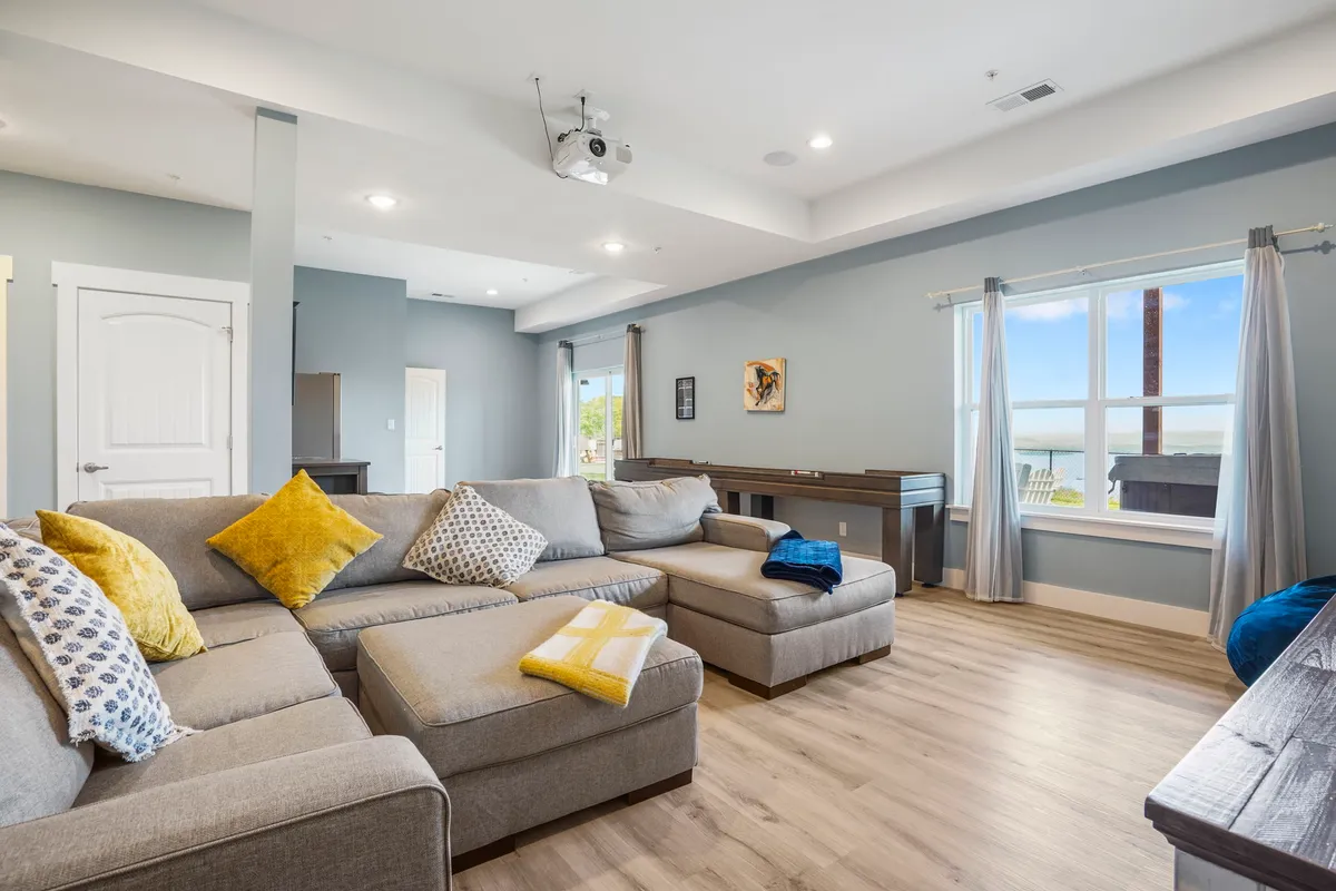 A spacious sectional sofa set beneath a ceiling-mounted projector, with large windows bringing in natural light and decorative throw pillows adding warmth. The arrangement invites viewers to envision themselves lounging for movies or competitive game nights.